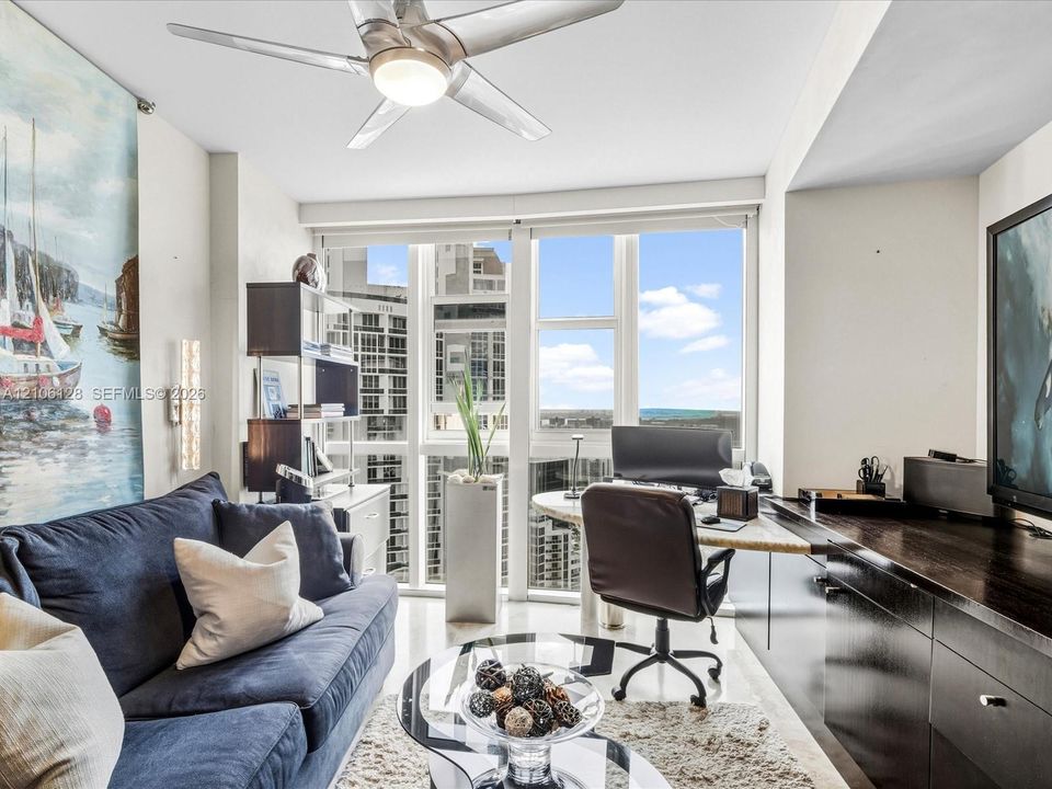Floor-to-ceiling windows, built-in dark cabinetry with integrated desk space, and a ceiling-mounted light fixture with fan