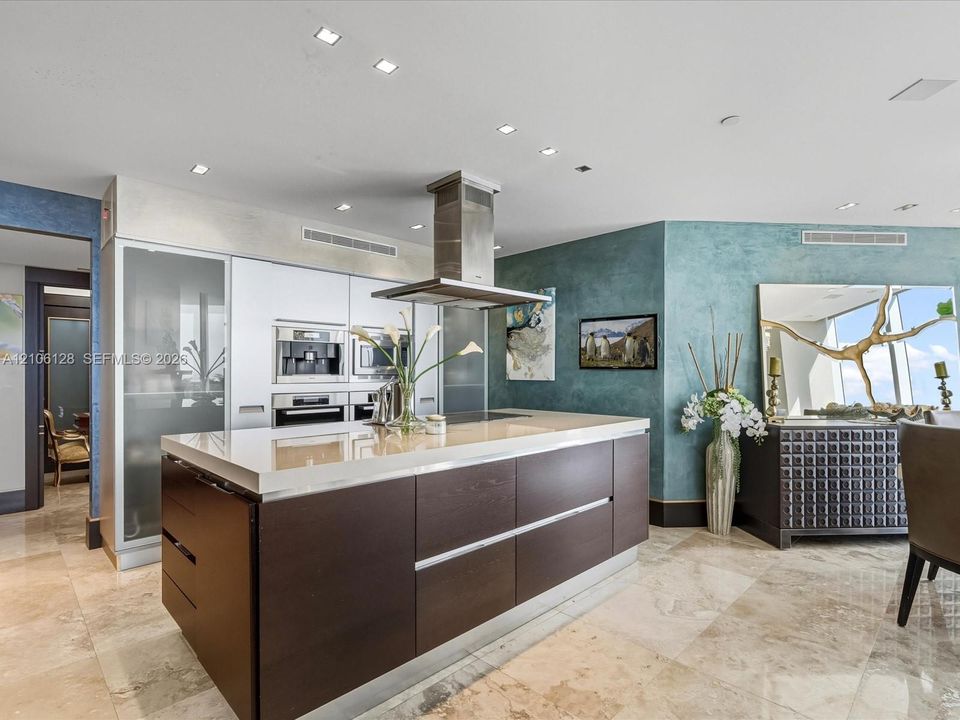Kitchen featuring a central island with a solid surface countertop, stainless steel overhead vent hood, built-in wall appliances, and stone flooring