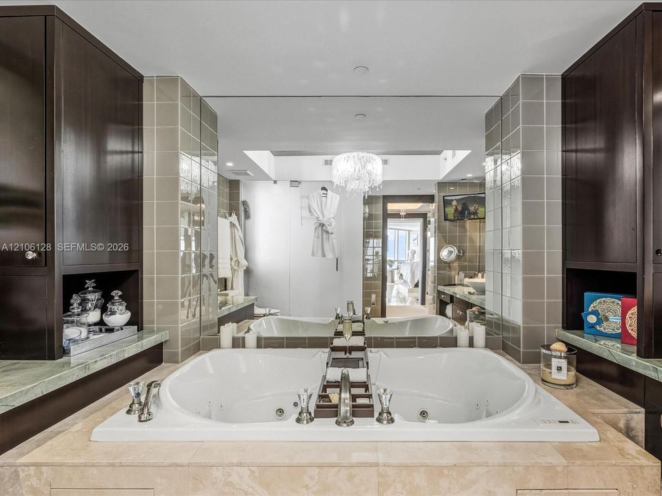Jetted soaking tub with deck-mounted chrome fixtures, flanked by dark wood cabinetry, integrated stone countertops, and full-wall mirror accented by tile surround and a central crystal-style chandelier