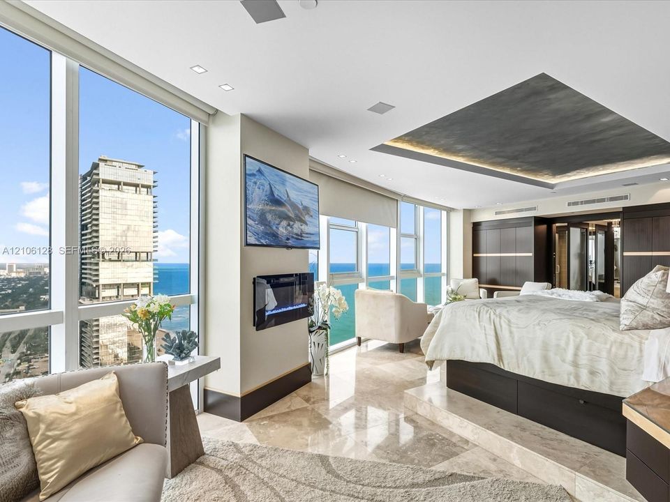 Floor-to-ceiling windows featuring ocean scenery, recessed ceiling detailing with integrated lighting, built-in wall-mounted electric fireplace, raised platform flooring with stone finish, and dark wood-finish built-in cabinetry