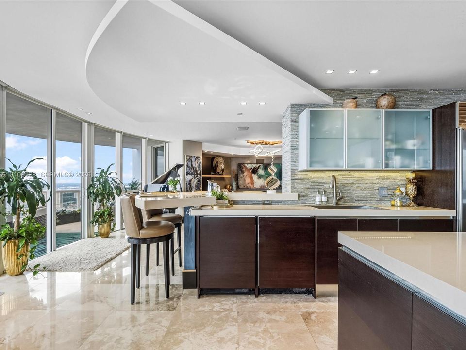 Gourmet kitchen featuring floor-to-ceiling glass walls, custom dark-finish cabinetry, stone-tile flooring, stacked-stone backsplash, and a curved architectural ceiling feature with recessed lighting