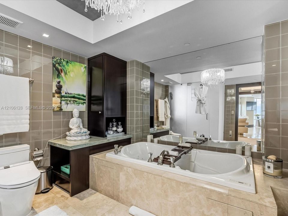 Bathroom featuring a stone-encased jetted soaking tub, dark-finish cabinetry with an integrated countertop, wall-to-wall mirror, tiled walls, and a crystal chandelier