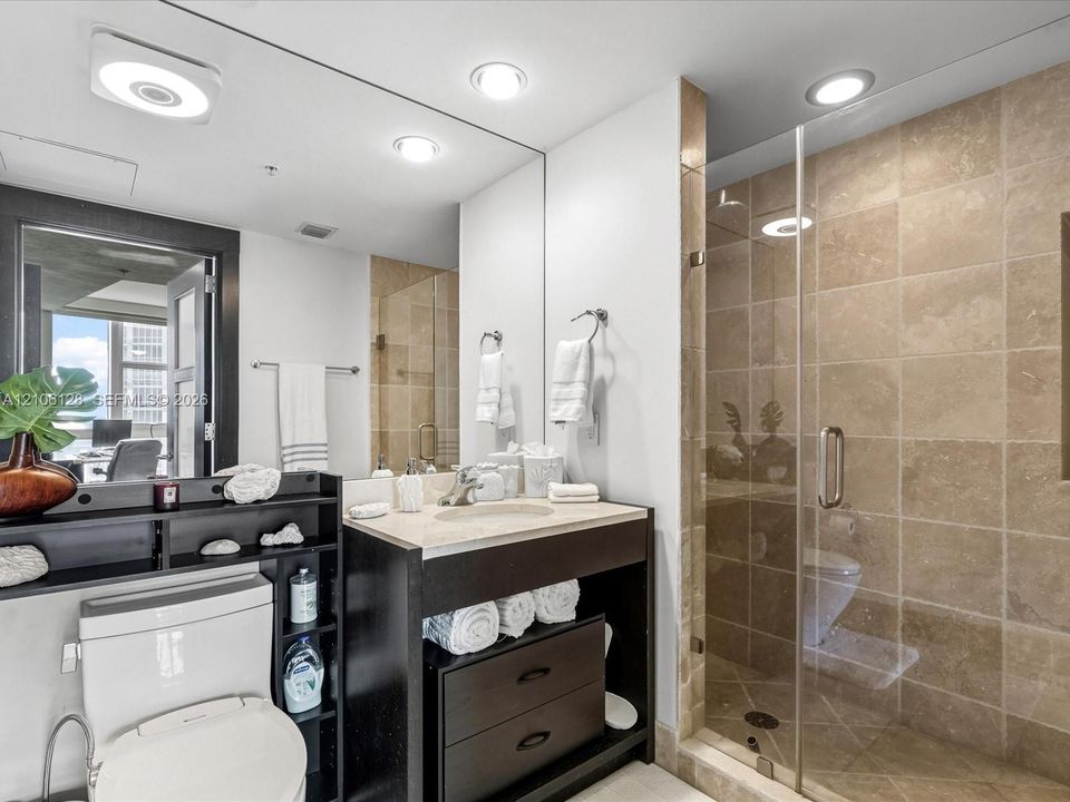 Bathroom featuring a vanity with stone countertop, under-mount sink, dark wood-finish cabinetry, full-width mirror, and a walk-in shower with tile surround and glass enclosure