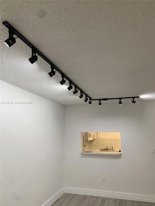 Ceiling-mounted matte black track lighting system featuring multiple adjustable spotlights, complemented by a service pass-through opening with a countertop ledge and wood-finish flooring