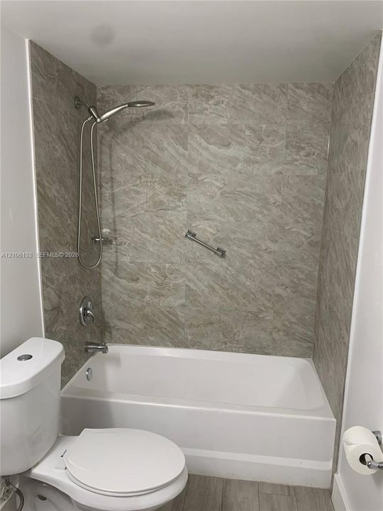 Full-size bathtub with stone-pattern tile surround, brushed nickel adjustable handheld shower fixture, built-in grab bar, and dual-flush toilet