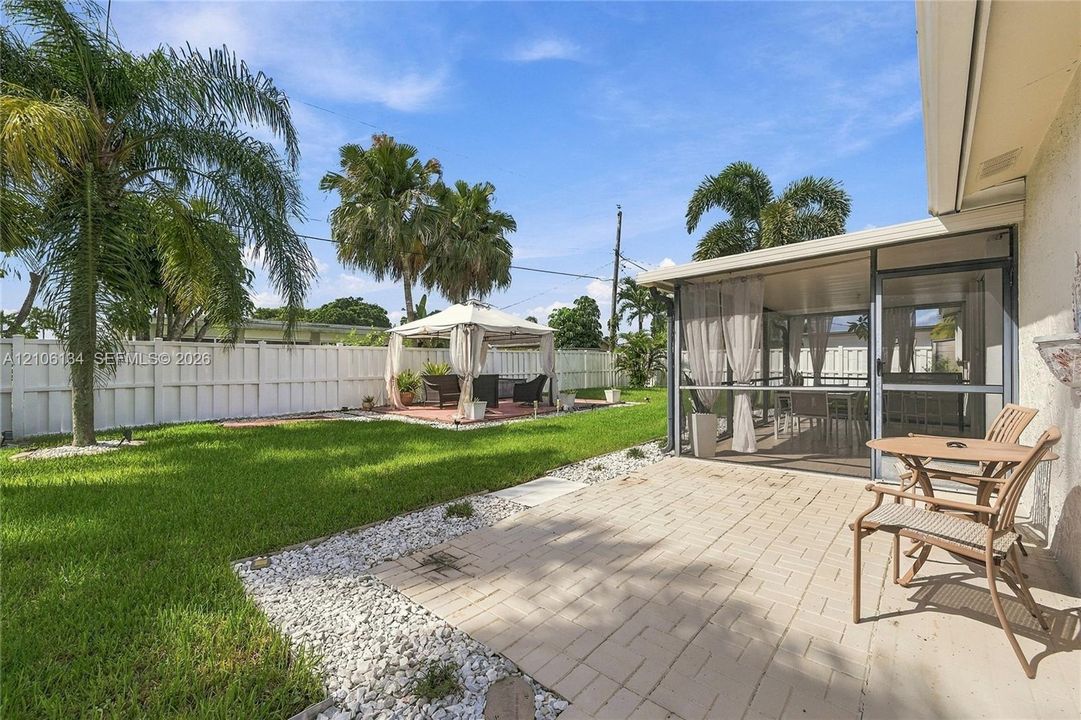 Backyard featuring a screened-in patio, concrete paver terrace, manicured lawn, white privacy fencing, and mature palm trees