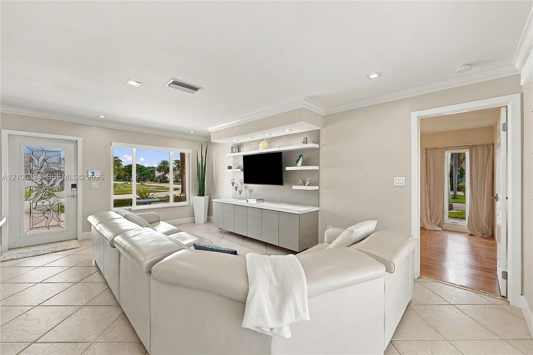 Living area featuring large-format diagonal tile flooring, custom built-in media cabinetry with integrated display shelving, crown molding, and a decorative glass-panel front door