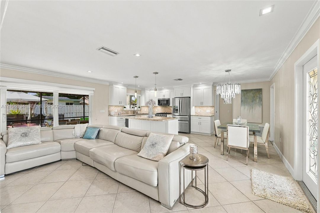 Open-concept interior featuring light-colored tile flooring, white shaker-style cabinetry, stone countertops, stainless steel appliances, and a tiered crystal chandelier
