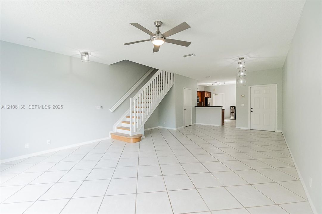 Open-concept living space featuring large-format diagonal tile flooring, a brushed-nickel ceiling fan with integrated lighting, a custom staircase with white vertical balusters and a wood-finished bottom tread, and modern multi-pendant lighting