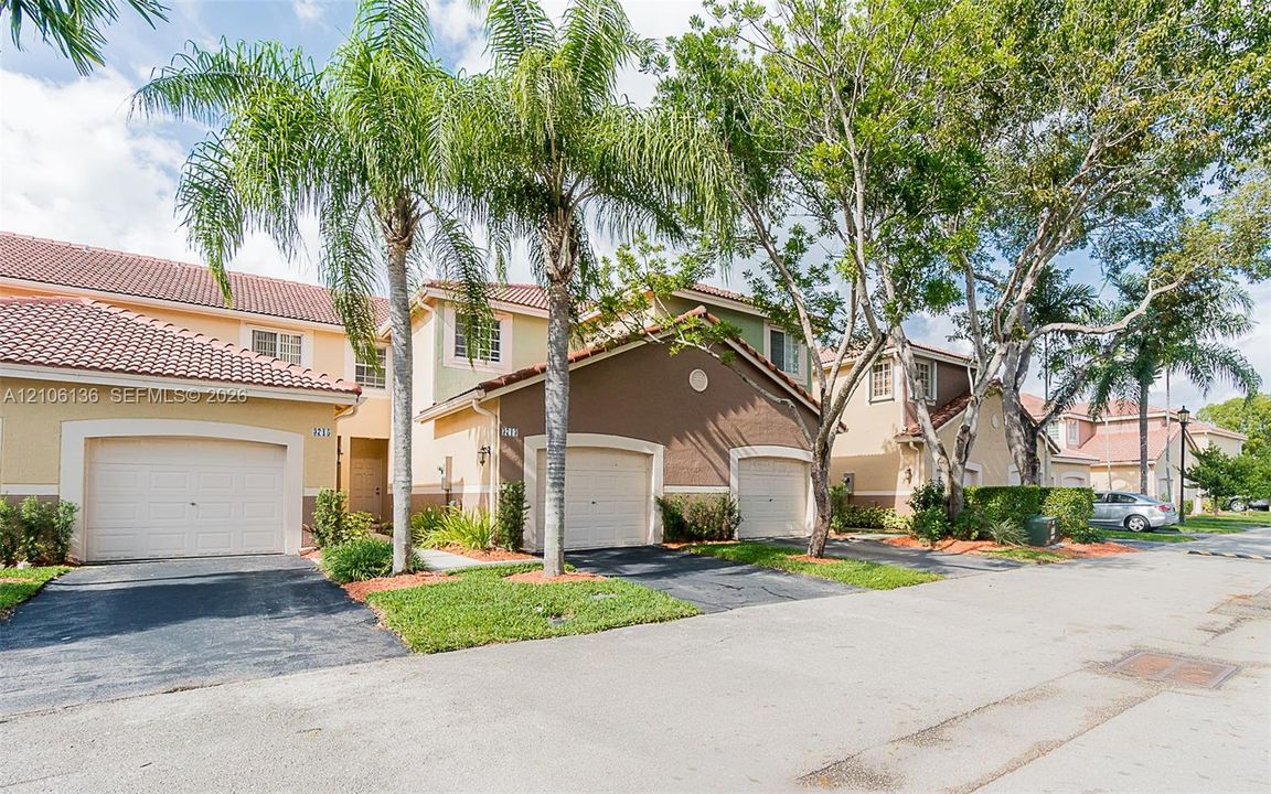 Multi-unit residential exterior featuring attached garages, tile roofing, stucco siding, paved driveways, and mature landscaping
