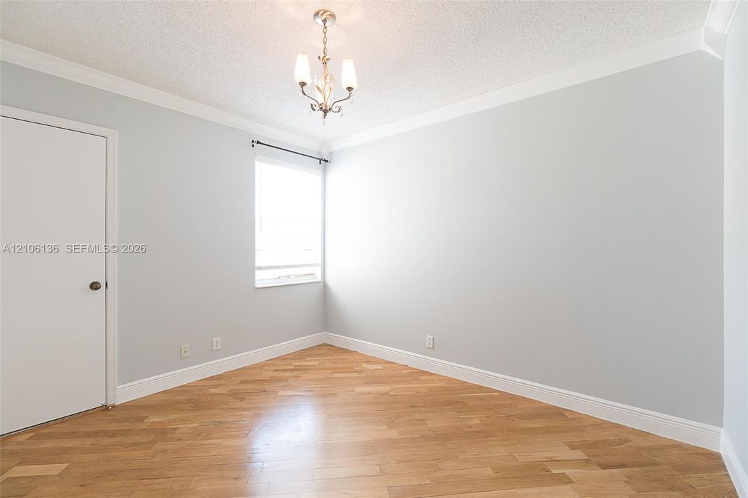 Bright corner bedroom with refinished wood-finish flooring, abundant natural light from large window, ceiling pendant fixture, and fresh white walls with crown molding detail