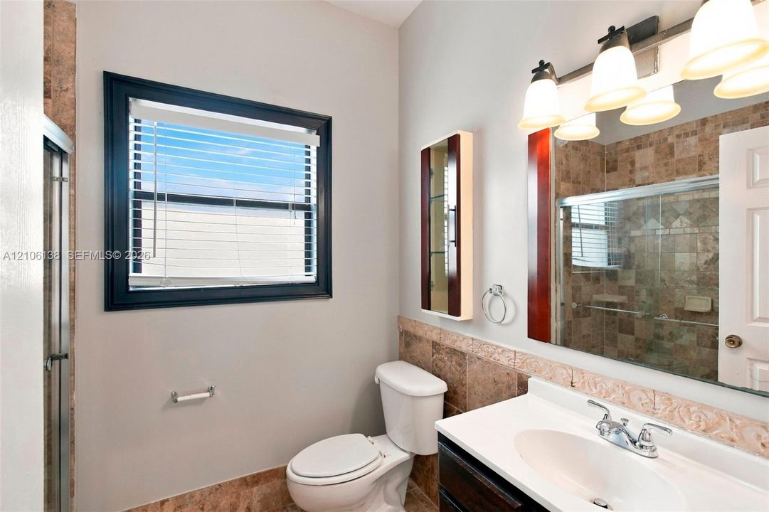 Bathroom featuring a single-sink vanity with a solid-surface countertop, tiled shower enclosure, stone-finish wall accents, and a large-format vanity mirror with integrated multi-bulb lighting