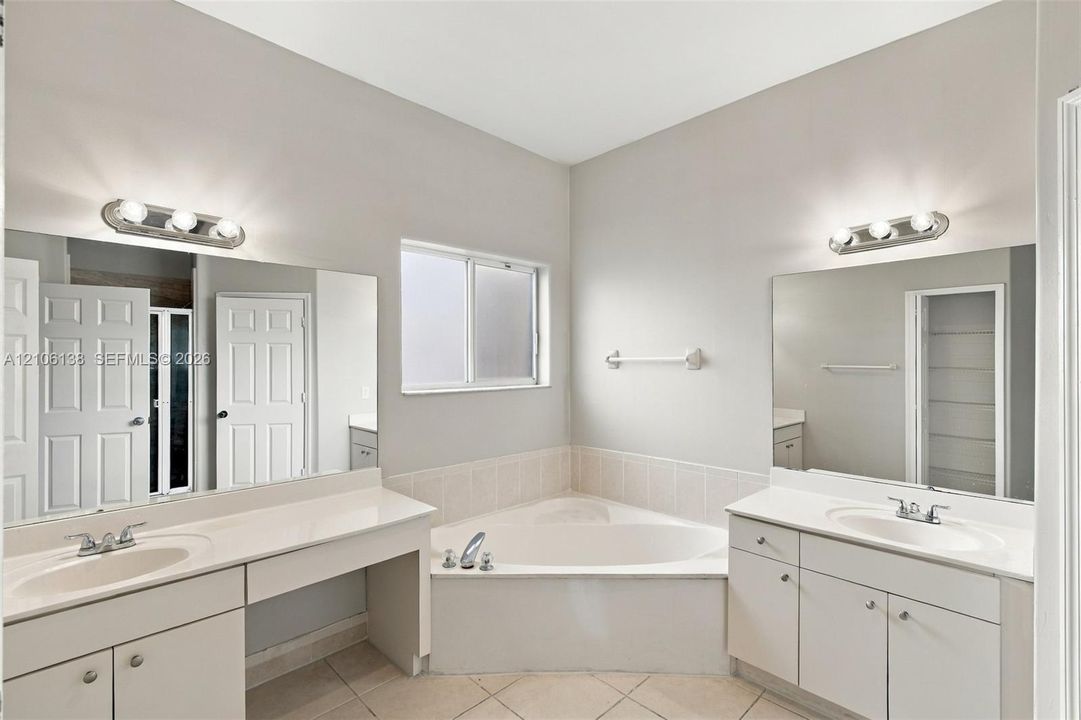Dual vanities with solid-surface countertops, tile-surround soaking tub, large wall-mounted mirrors, and multi-bulb light fixtures