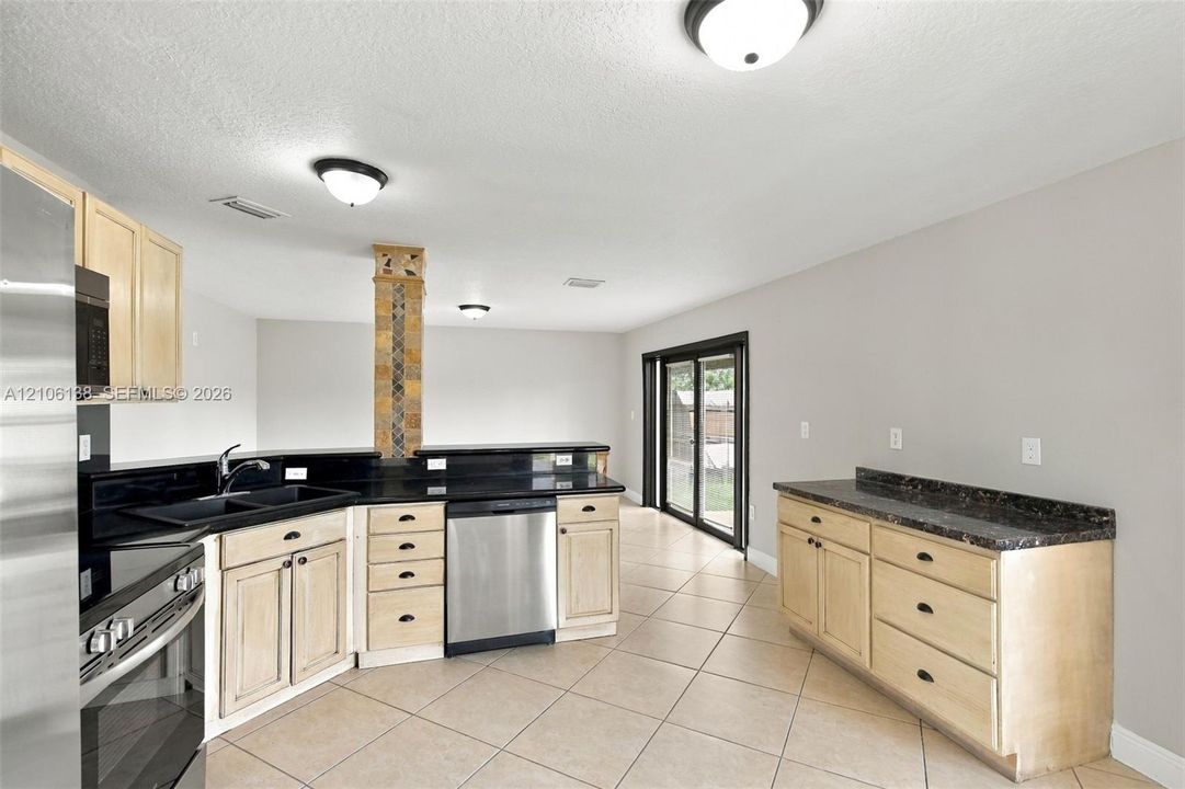 Kitchen featuring light wood-finish cabinetry, dark solid-surface countertops, built-in stainless steel appliances, a decorative tiled support pillar, and ceramic tile flooring