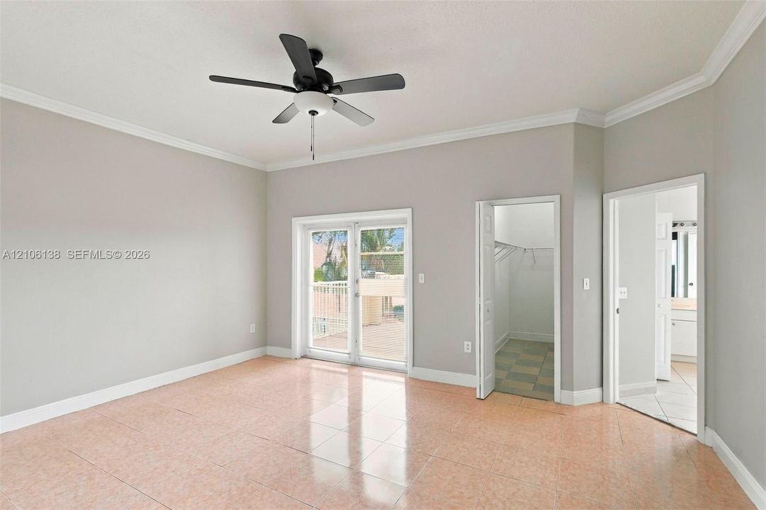 Neutral-toned room featuring tile flooring, crown molding, a ceiling fan with integrated light fixture, glass-paned double doors, and entryways to a walk-in closet and bathroom