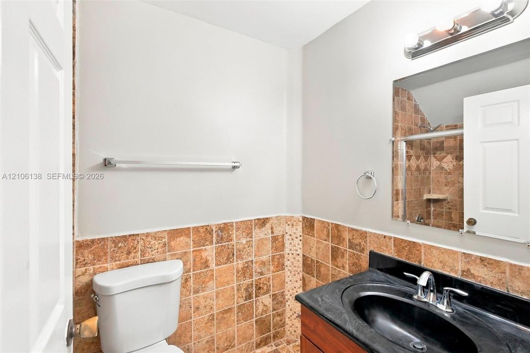 Bathroom featuring a wood-tone vanity, dark solid-surface countertop with an integrated oval sink, mid-tone stone tile wainscoting, and a wall-mounted light fixture