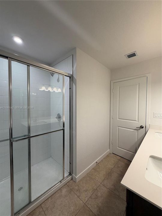 Walk-in shower with chrome-framed glass enclosure and tiled surround, dual-sink vanity with solid-surface countertop, and ceramic tile flooring