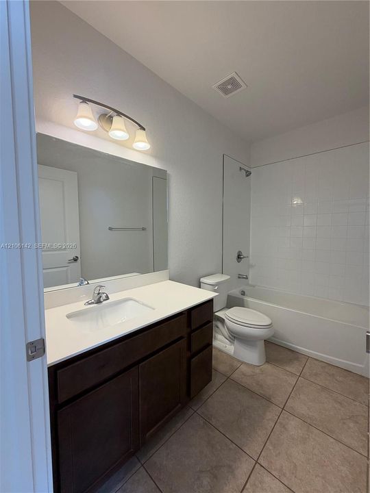 Dark wood-finish cabinetry, white countertop with integrated sink, large wall-mounted mirror, three-light vanity fixture, and tiled tub-shower surround
