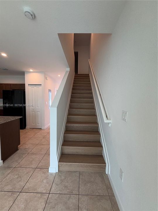 Staircase featuring wood-finish treads, a white handrail, and ceramic tile flooring throughout the main level