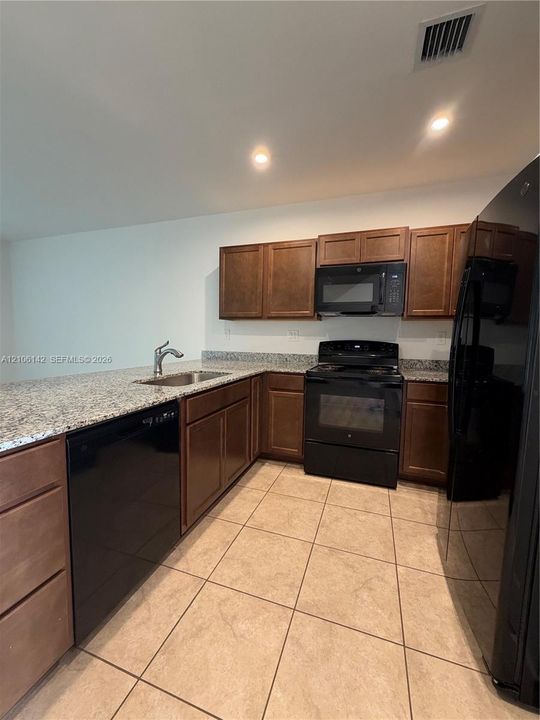 Kitchen featuring medium-toned wood cabinetry, light-colored stone-finish countertops, tile flooring, and integrated black appliances