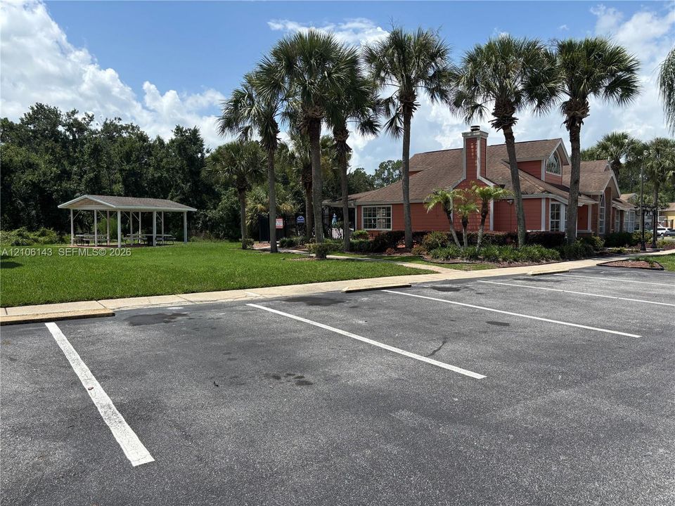 Asphalt parking area featuring marked spaces, a manicured lawn, a covered pavilion, and mature palm trees