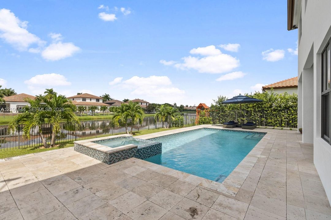Outdoor living area featuring a paved stone deck, in-ground swimming pool, raised spill-over spa with decorative tile inlay, and a metal perimeter fence