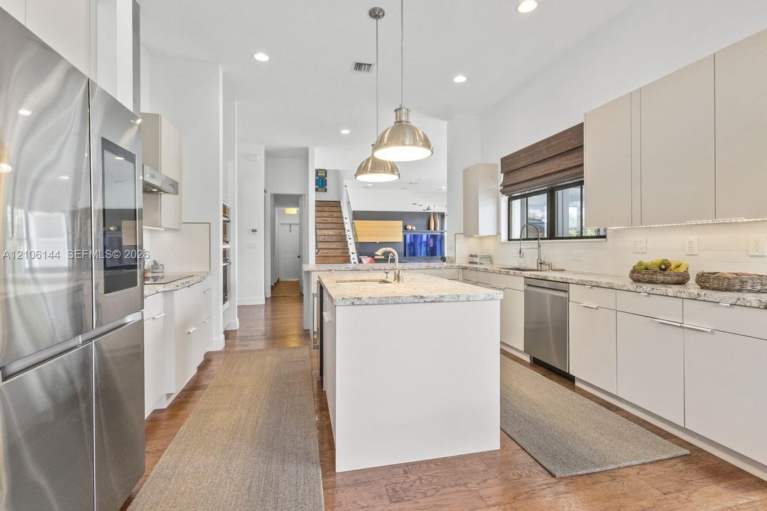 Kitchen featuring a central island with a stone-finish countertop, stainless steel appliances, flat-panel cabinetry, wood-finish flooring, and industrial-style pendant lighting