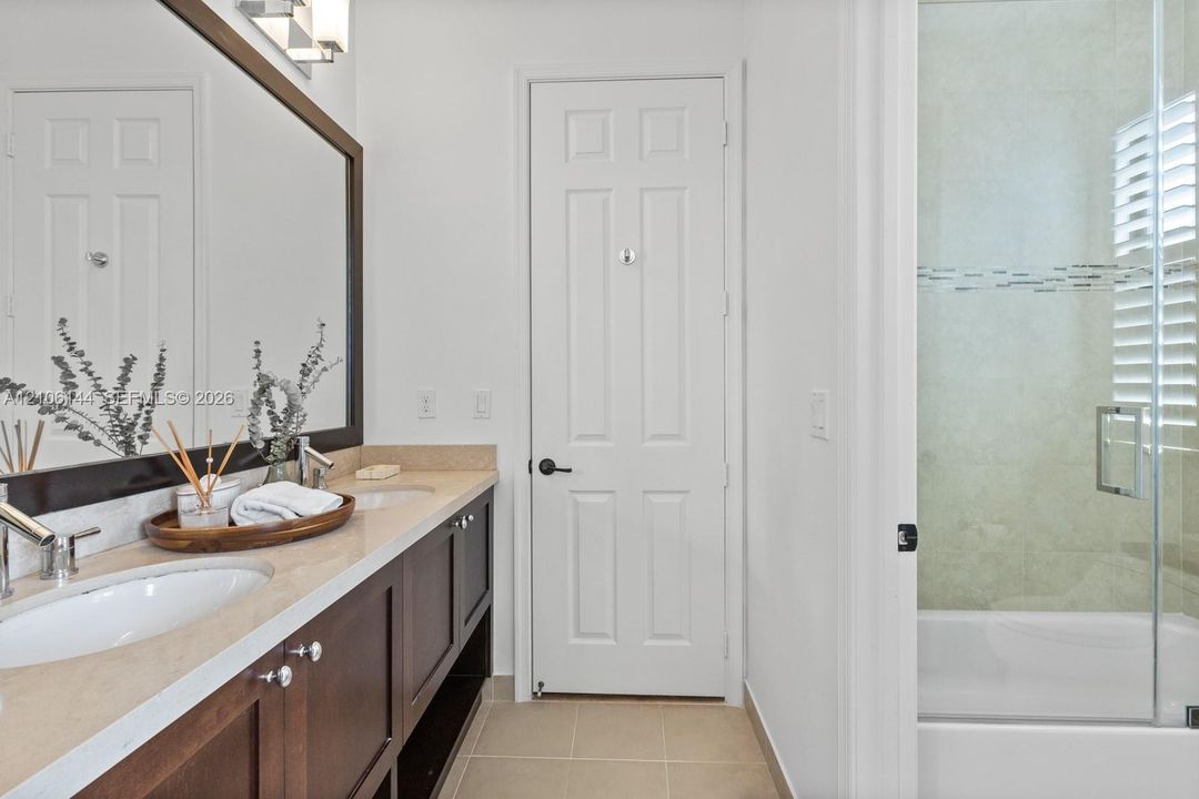 Dual-sink vanity with stone-finish countertop, dark wood cabinetry, full-width mirror, and glass-enclosed shower with tile surround