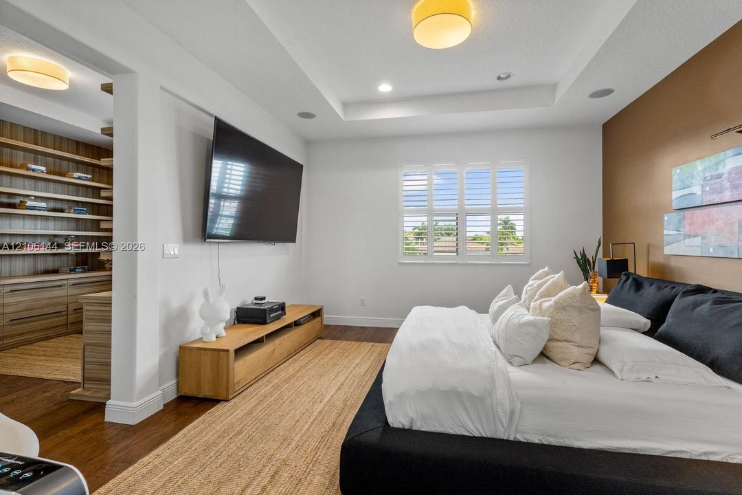 Tray ceiling with recessed lighting, plantation shutters, built-in shelving, wood-finish flooring, and wall-mounted light fixtures