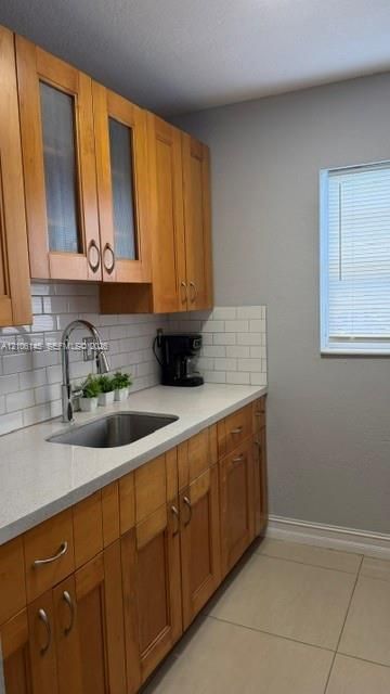Natural wood cabinetry with glass-paneled upper doors, white subway tile backsplash, light-toned stone-finish countertops, stainless steel undermount sink with high-arc faucet, and large-format tile flooring