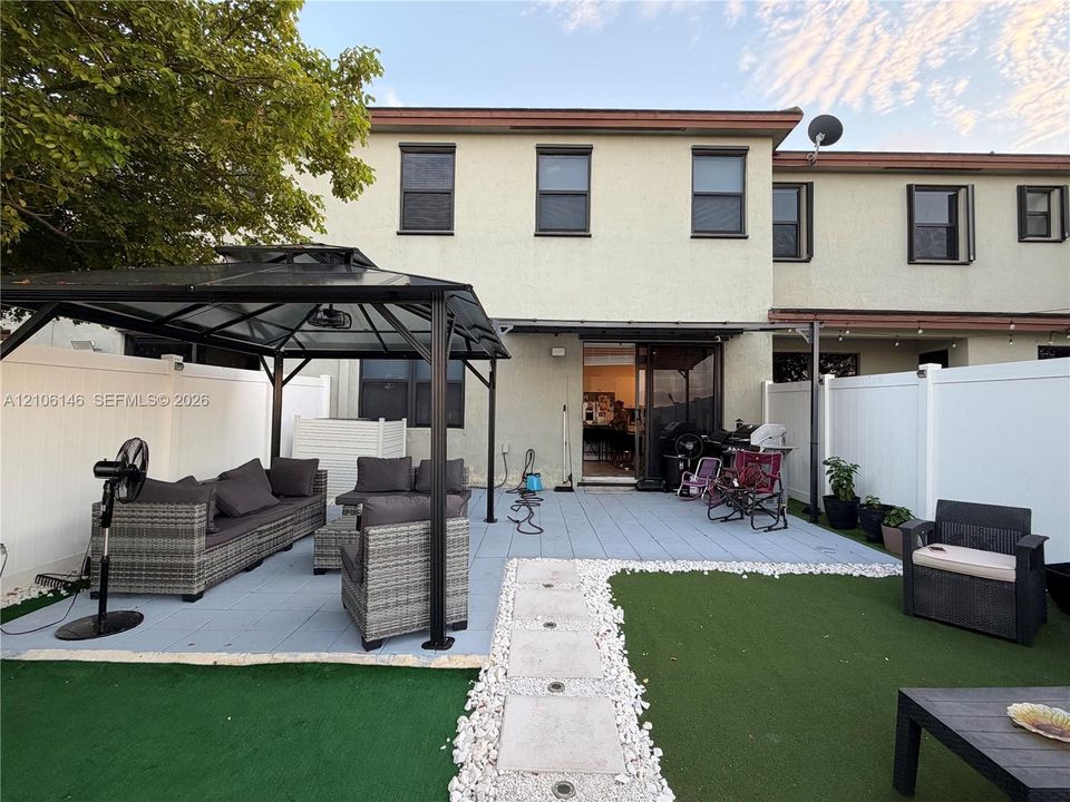 Backyard featuring a paved patio, synthetic turf lawn, white vinyl perimeter fencing, and stone-bordered walkway