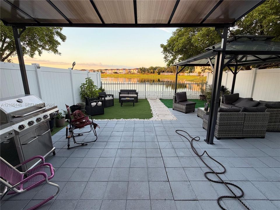 Paved patio featuring a metal gazebo, synthetic turf lawn, white vinyl fencing, and lake frontage
