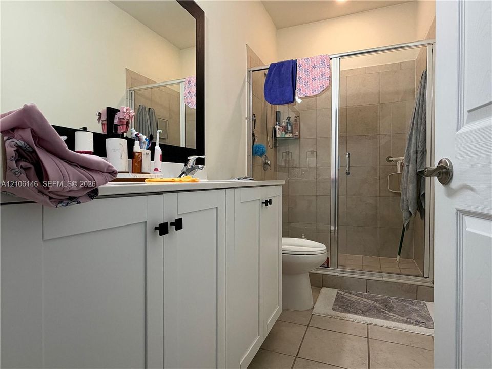 Bathroom featuring a white shaker-style vanity with matte black hardware, a large framed mirror, tiled flooring, and a walk-in shower with a glass enclosure and tiled surround