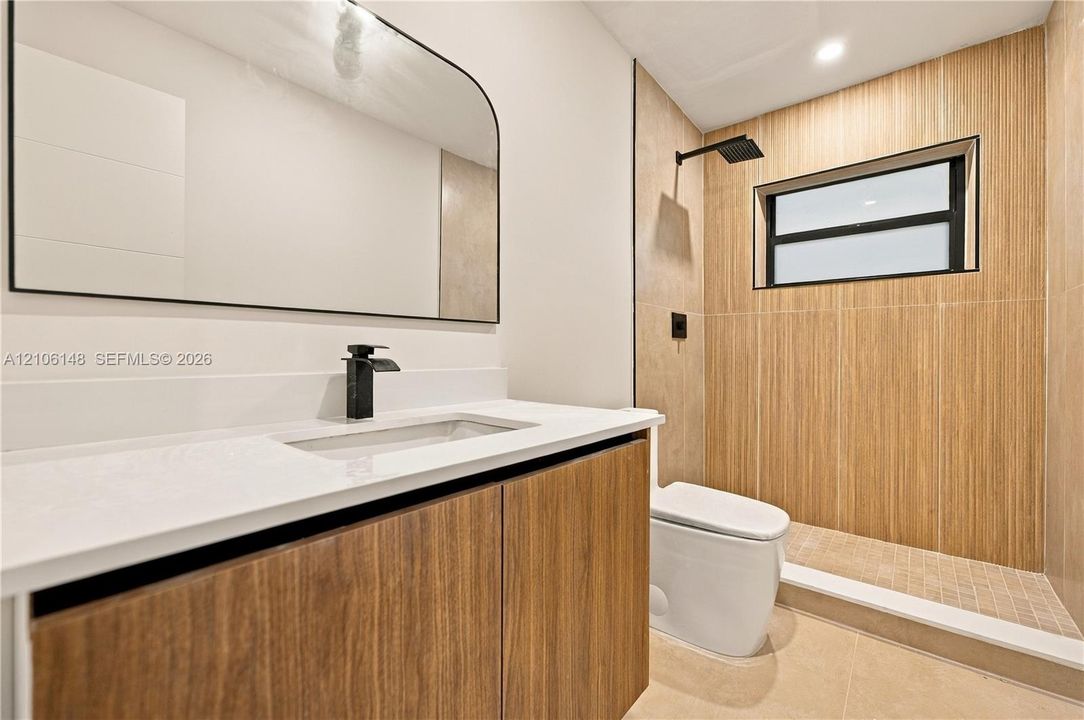Floating vanity with wood-finish cabinetry, white countertop, arched mirror, matte black fixtures, and walk-in shower with vertical-tiled surround