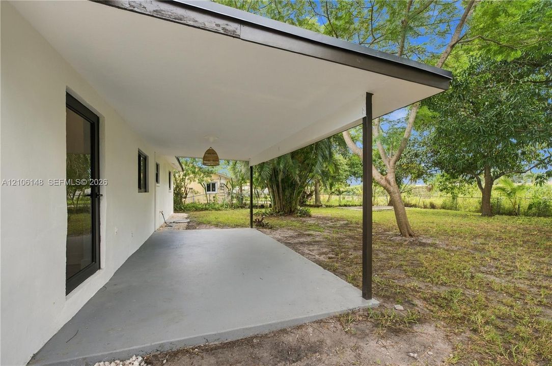 Covered concrete patio featuring a solid support post, recessed ceiling lighting, and exterior access door, set against a spacious yard with mature trees