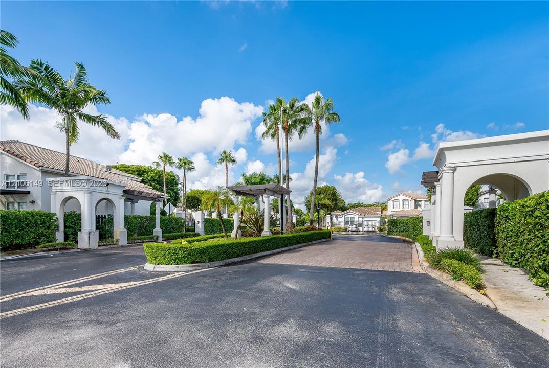 Gated entrance featuring decorative stone pillars, arched stucco monuments, paver driveway surfacing, and manicured perimeter hedging