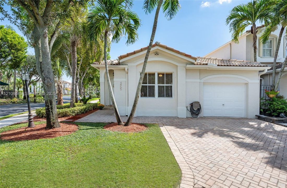 Stucco exterior with a tiled roof, arched entryway, brick-paver driveway, single-car garage, and established palm tree landscaping