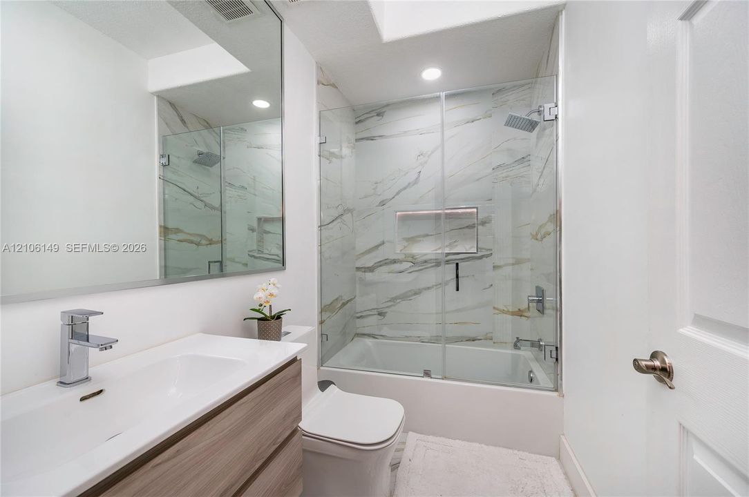 Bathroom featuring a wood-finish floating vanity with a white rectangular sink, a large wall-mounted mirror, a tub-shower combination with floor-to-ceiling stone-patterned wall tile, a glass shower enclosure, and a built-in recessed wall niche