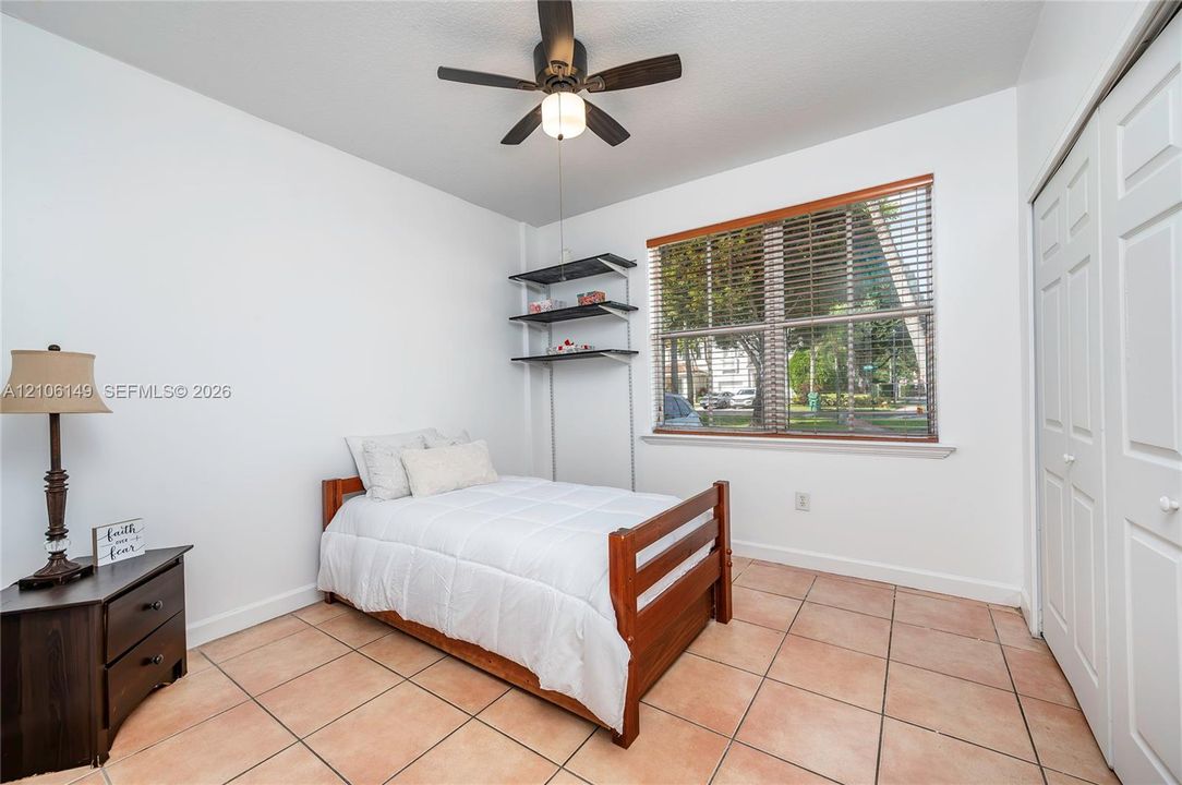 Bedroom featuring tile flooring, ceiling fan with integrated lighting, window with horizontal blinds, built-in shelving, and dual-panel closet doors