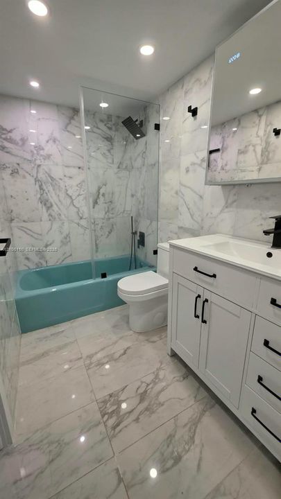 Full-height tile walls with a veined pattern, glass shower enclosure, white vanity cabinet with matte black hardware, integrated rectangular sink, and recessed ceiling lighting