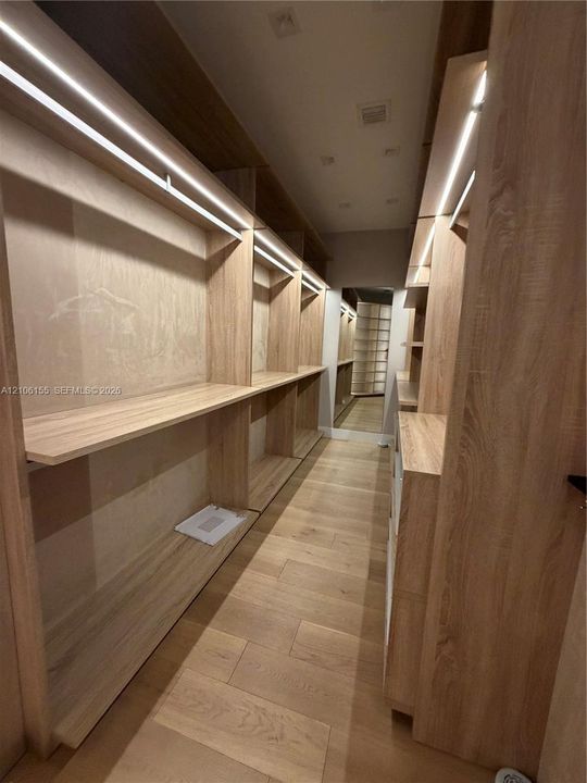Custom Men’s Primary closet w storage system featuring light wood-finish cabinetry, integrated linear LED shelving illumination, open-concept shelving units, and wood-finish flooring
