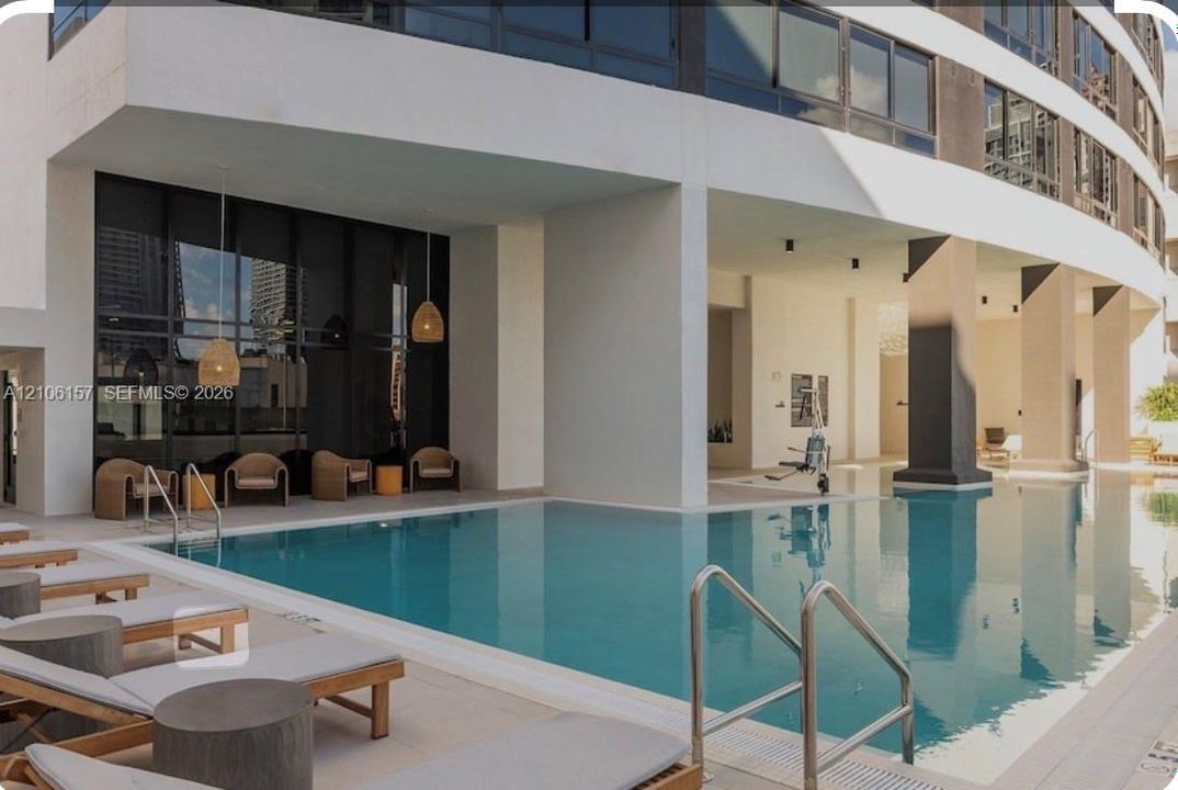Outdoor swimming pool featuring a concrete deck, dual metal handrails, and a covered architectural overhang with recessed lighting