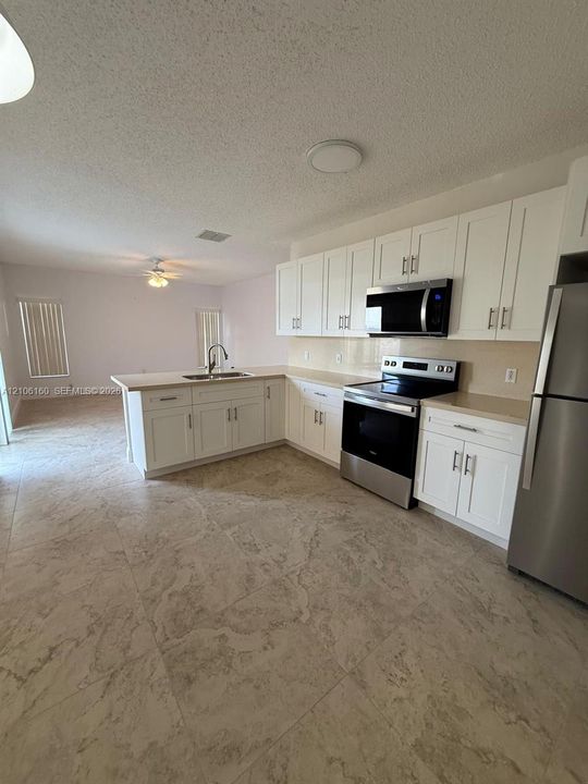 Kitchen featuring white Shaker-style cabinetry, stainless steel appliances, neutral-toned stone-finish tile flooring, a peninsula with a double-basin sink, and solid-surface countertops
