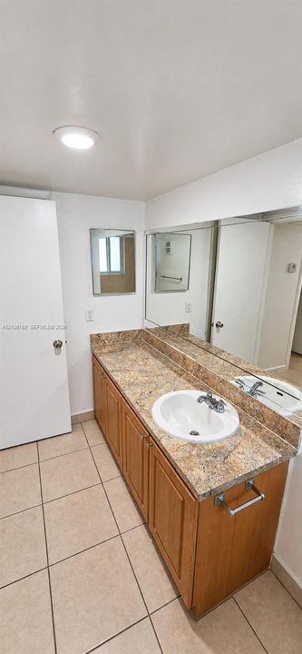 Vanity featuring wood-finish cabinetry, a stone-patterned countertop, an integrated circular sink, a full-width wall mirror, and neutral floor tiling