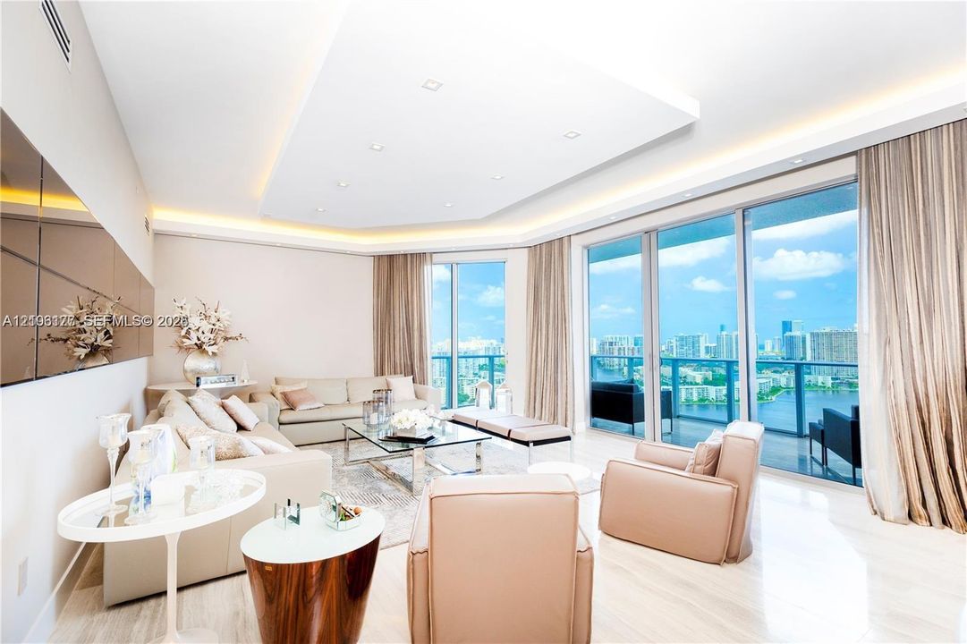 Living area featuring floor-to-ceiling glass panel walls, tray ceiling with recessed perimeter lighting, light-toned polished flooring, and a glass-paneled balcony