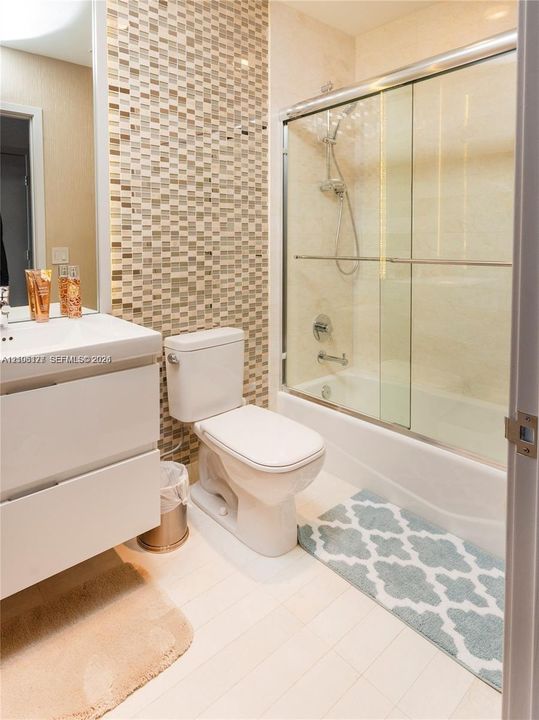 Bathroom featuring a white floating vanity, mosaic tile accent wall, and a tub-shower combination with a glass sliding enclosure and handheld shower fixture
