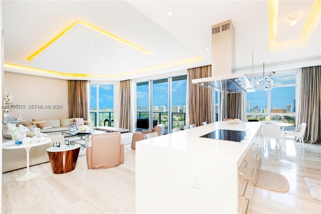 Open-concept living space featuring a central kitchen island with a built-in cooktop, stainless steel vent hood, recessed tray ceilings with perimeter lighting, and floor-to-ceiling glass windows