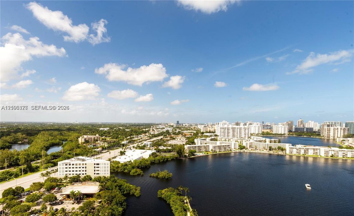 Panoramic aerial perspective featuring expansive lake frontage, lush tropical vegetation, mid-rise developments, and a distant cityscape horizon