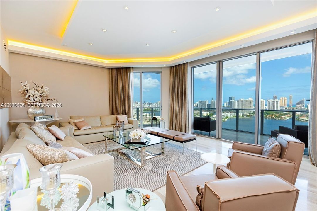 Floor-to-ceiling glass sliding doors, recessed tray ceiling with integrated cove lighting, polished neutral-toned flooring, and expansive panoramic water and cityscape horizon