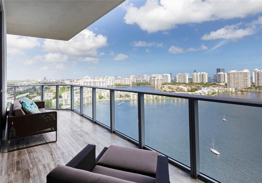 Private balcony with stone-finish flooring and floor-to-ceiling glass railings overlooking the waterway and city skyline
